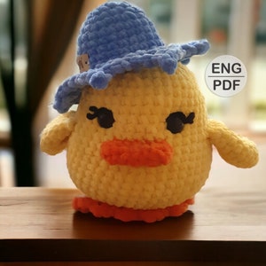 May include: A crocheted yellow chick wearing a blue crocheted hat with a wide brim. The chick has black eyes, a small orange beak, and orange feet. The image includes the text "ENG PDF".