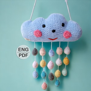 Pode incluir: Uma nuvem azul de crochê com um rosto sorridente e bochechas cor-de-rosa pendura em um cordão branco.  Presos à nuvem, há pompons de crochê coloridos em tons de azul, rosa, amarelo e branco. O texto "ENG PDF" está impresso em um círculo branco.