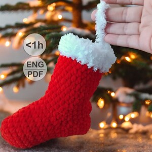 May include: Red crocheted Christmas stocking with a white faux fur cuff and a loop at the top. The stocking is hanging from a hand. The text '<1h ENG PDF' is visible in the image.
