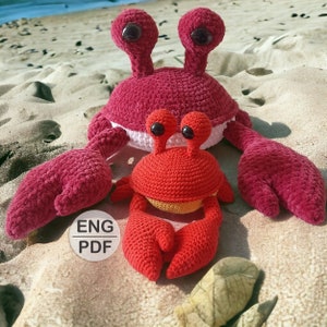 Puede incluir: Dos cangrejos de peluche tejidos a crochet, uno grande y otro pequeño. El cangrejo más grande es rojo con detalles blancos y el cangrejo más pequeño es rojo con detalles amarillos. Ambos cangrejos tienen ojos negros y están sentados en una playa de arena.