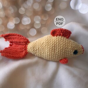 May include: A crocheted goldfish toy with a cream-colored body and orange fins and tail. The toy has a black eye and is lying on a white surface.