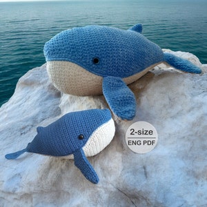 May include: Two crocheted whale plushies, one large and one small, are sitting on a white rock. The whales are blue with white bellies and black eyes. The larger whale is lying down with its tail curled up. The smaller whale is sitting up with its tail curled under its body. A white circle with the text "2-size ENG PDF" is in the lower right corner of the image.