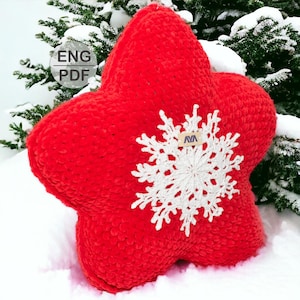 May include: A red crocheted star-shaped pillow with a white crocheted snowflake design in the center. The pillow is sitting on a snowy surface.
