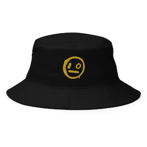 May include: Black bucket hat with a yellow embroidered smiley face design. The hat features a wide brim and a classic bucket hat silhouette. The yellow smiley face has a unique, slightly distressed look.