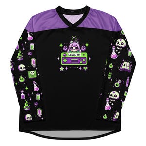 Puede incluir: Jersey de manga larga negro y morado con un gráfico de mando de videojuego verde y morado y un personaje de dibujos animados con el texto "LEVEL UP". Las mangas presentan un patrón repetido de frascos de pociones verdes y morados, calaveras y otros objetos mágicos.