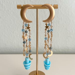 May include: Gold-coloured crescent moon earrings with dangling chains of stars, beads, and small moons. The earrings feature light blue earplugs at the bottom. The earrings are displayed on a gold stand.