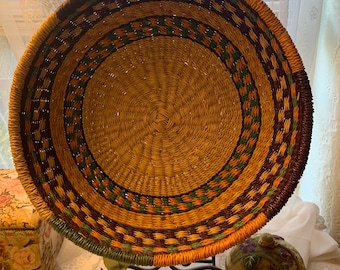 May include: A round, woven basket with a decorative pattern in shades of brown, green, and orange. The basket is displayed on a black metal stand. The basket's interior is a light tan color.