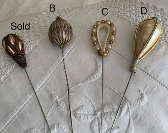May include: Four decorative hat pins labeled Sold, B, C, and D. The first is brown with metallic accents, B is a silver filigree sphere, C features a loop of cream pearls, and D is a smooth cream teardrop shape with silver wire wrapping.