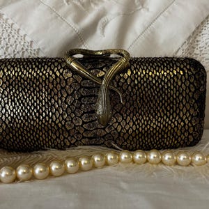 May include: A rectangular clutch bag featuring a black and metallic gold snakeskin pattern. The closure is a prominent brass-toned metal ornament shaped like a coiled snake. A strand of cream-colored faux pearls is positioned in front of the bag.