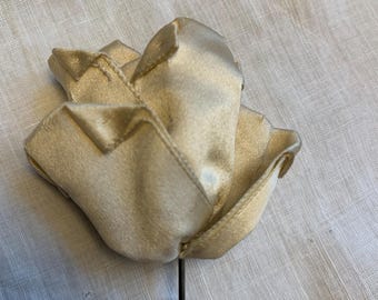 May include: A cream-colored fabric rose with detailed petal construction. The flower is attached to a thin, dark stem. The rose is set against a light-colored, textured background.