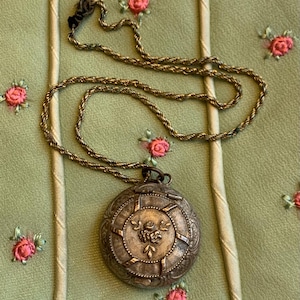 May include: Antique silver-toned pendant necklace with a detailed, round locket. The locket features a floral design at the center and is suspended from a delicate chain. The background is a light green fabric with embroidered pink roses.