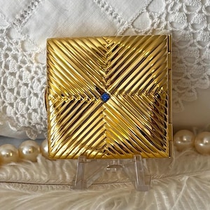 May include: A square gold-toned metal compact case featuring a diagonal ribbed texture that converges toward the center. A small, square blue stone is embedded in the middle of the design. The compact has a hinged side and a metallic finish.