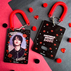 Op de afbeelding: Twee zwarte ID-badgehouders met rode spiraalsleutelhangers. Eén heeft een foto van een persoon en het woord "BADDIE". De andere zegt "EMOTIONAL SUPPORT BADDIE". Beide hebben rode lippen en hartontwerpen.
