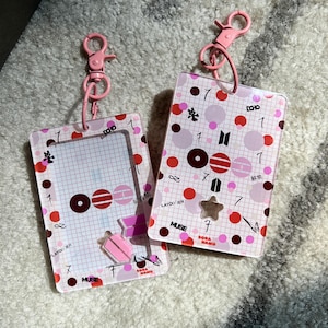 May include: Two clear acrylic keychains with pink hardware. The keychains have a grid pattern with red, pink, and brown circles and shapes. Text includes "DCHO", "MUSE", and "BORA HAMU".
