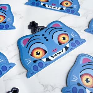 May include: A collection of blue cat-shaped stickers with cartoonish features. Each sticker has large orange eyes, white teeth, and pink accents. A small black sticker with a hat is also present.