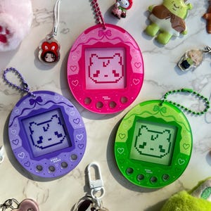 Puede incluir: Tres llaveros de mascotas digitales coloridos en rosa, morado y verde, cada uno con un diseño de oso pixelado en la pantalla. Otros amuletos incluyen una figura de Shrek y un amuleto en forma de corazón de BTS.
