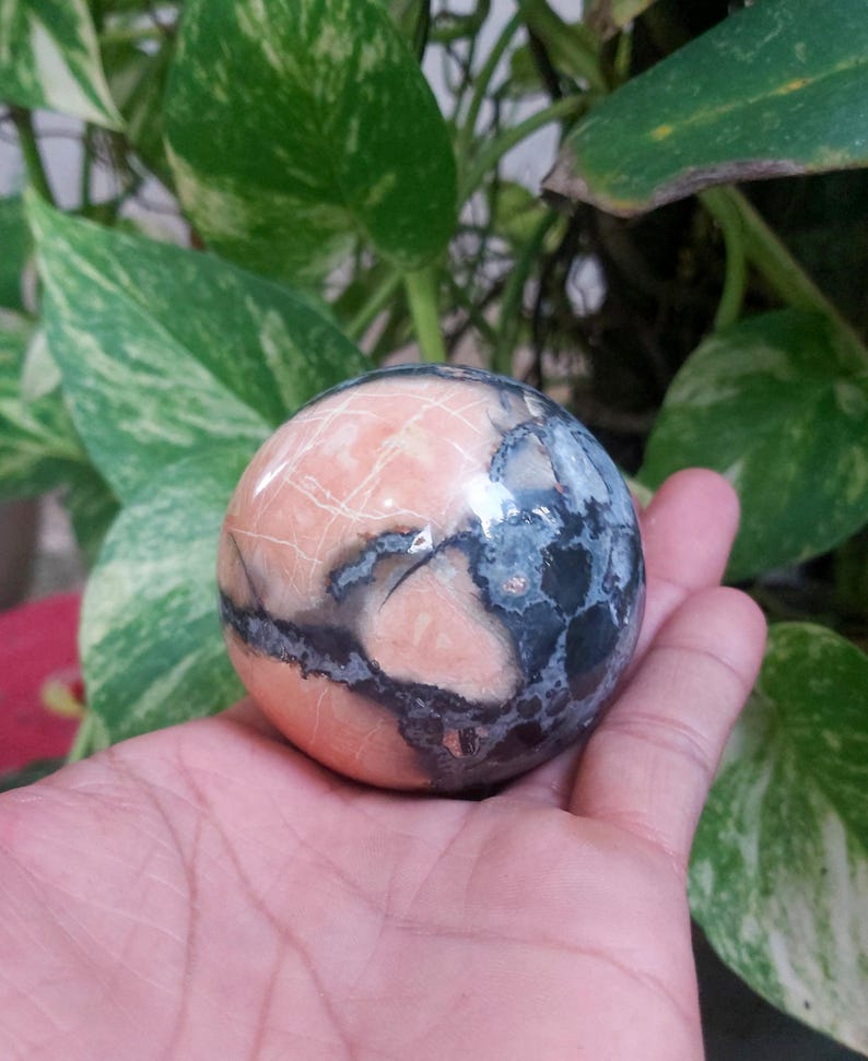 May include: A polished sphere of pink and gray stone with a swirling pattern. The sphere is smooth and has a high gloss finish.