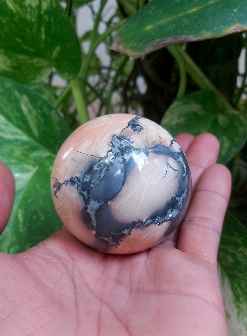 May include: A smooth, polished sphere of pink and gray stone with intricate patterns. The sphere is about the size of a golf ball and has a high gloss finish.