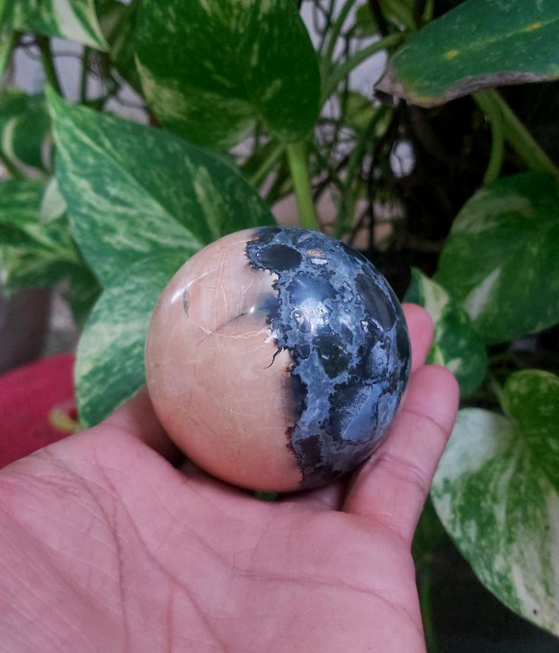 May include: A smooth, round, polished stone sphere with a half-and-half design. One half is a light pink color, and the other half is a dark blue with white speckles.