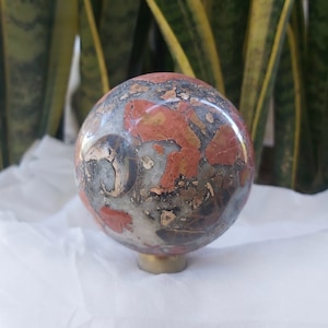 Op de afbeelding: Een ronde, gepolijste bol van rode, zwarte en witte jaspis met een messing voet. De bol heeft een draaiende, gemarmerde structuur.