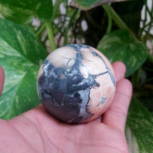May include: A polished sphere of pink, gray, and white ocean jasper. The sphere has a swirling pattern of colors and is smooth to the touch.