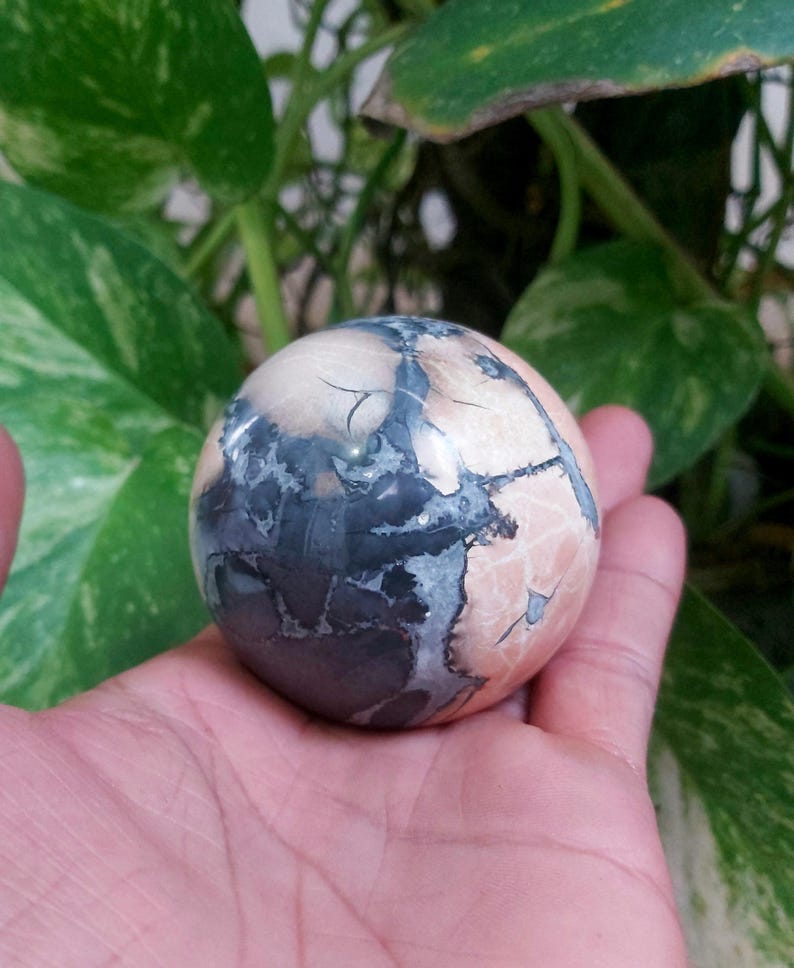 May include: A polished sphere of pink, gray, and white ocean jasper. The sphere has a swirling pattern of colors and is smooth to the touch.