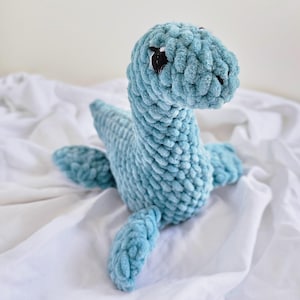 May include: A blue crocheted plush toy shaped like a dinosaur. The toy has large black eyes and a long neck.