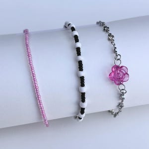 May include: Three bracelets are displayed against a white background. One is a thin strand of pink beads. Another features alternating black and white beads. The third has a silver chain with a pink rose charm.