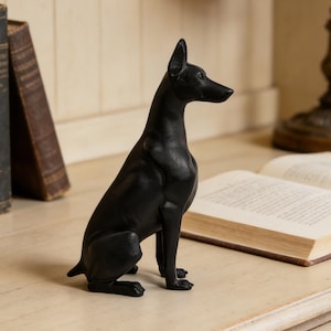 May include: A black dog figurine in a seated position. The statue has a sleek, glossy finish and is positioned on a light-colored wooden surface. The dog's ears are perked up, and it is facing to the right. Books and a lamp are in the background.