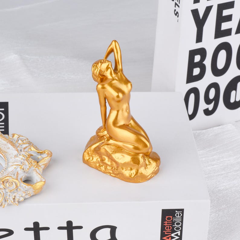 May include: A gold-colored figurine of a reclining nude figure, posed with one arm raised. The sculpture is mounted on a textured base. The figurine is a decorative item, likely for home decor.