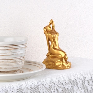 May include: A gold-colored mermaid statue, posed on a rock, with one arm raised above her head. The statue is placed on a white lace tablecloth next to a white teacup and saucer with brown stripes.