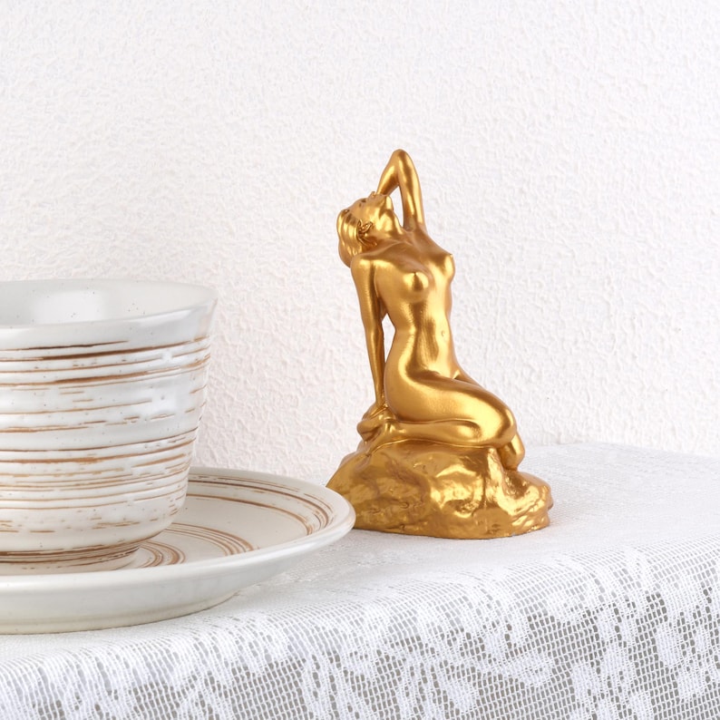 May include: A gold-colored mermaid statue, posed on a rock, with one arm raised above her head. The statue is placed on a white lace tablecloth next to a white teacup and saucer with brown stripes.