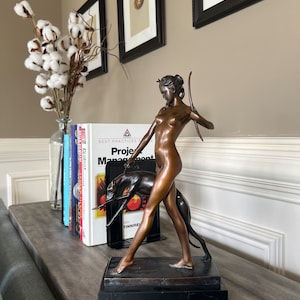 May include: A bronze-toned sculpture of a figure holding a bow and walking with a dog, mounted on a rectangular black base. The figure is depicted in a standing pose with one arm raised, while the dog is positioned beside the figure's legs.