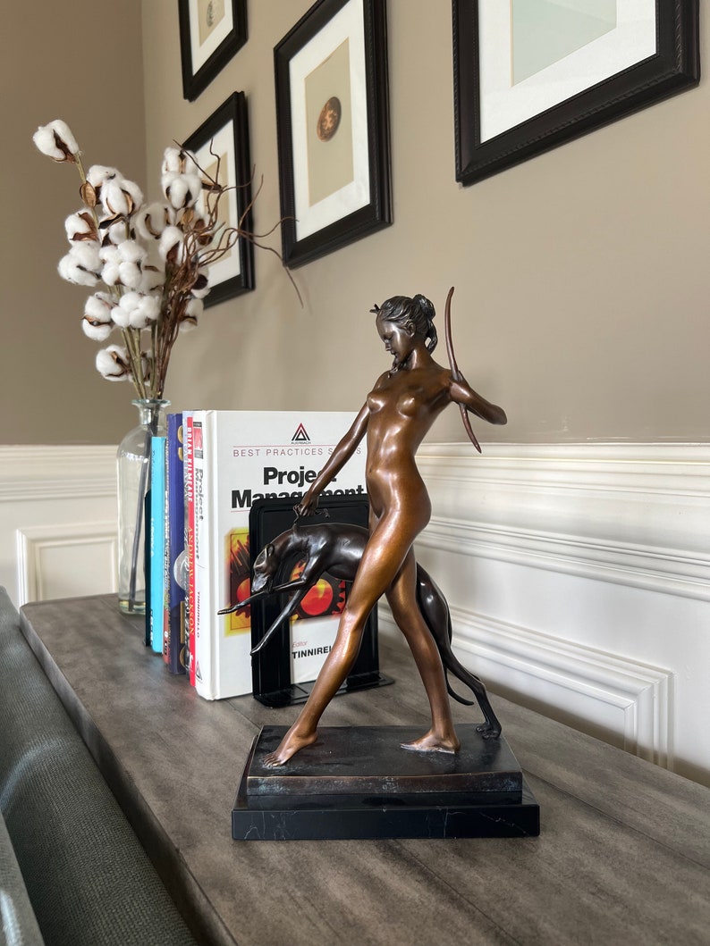 May include: A bronze-toned sculpture of a figure holding a bow and walking with a dog, mounted on a rectangular black base. The figure is depicted in a standing pose with one arm raised, while the dog is positioned beside the figure's legs.