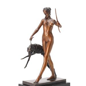 May include: A bronze-toned sculpture of a standing figure holding a bow in one hand and a leash attached to a dark-colored dog in the other. The figure is depicted in a walking pose, mounted on a rectangular black base with a smooth, polished finish.