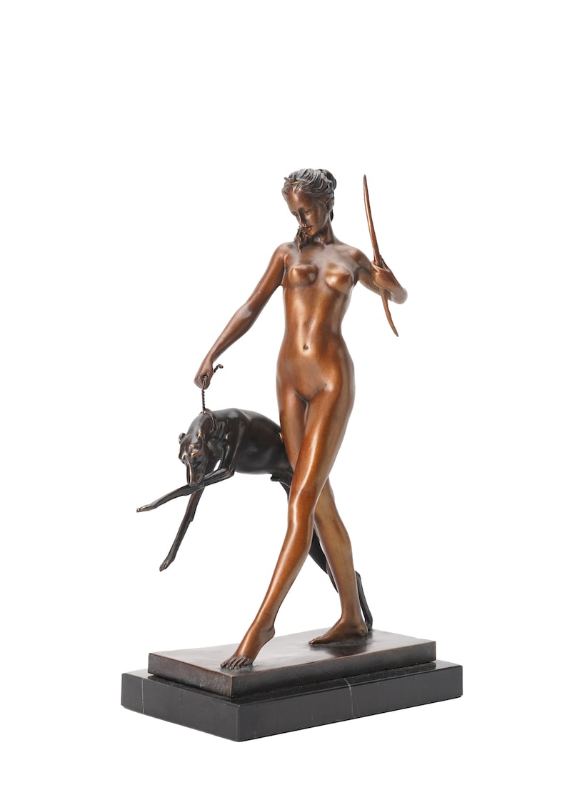 May include: A bronze-toned sculpture of a standing figure holding a bow in one hand and a leash attached to a dark-colored dog in the other. The figure is depicted in a walking pose, mounted on a rectangular black base with a smooth, polished finish.