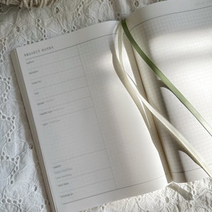 Könnte beinhalten: Ein offenes Projektplaner-Notizbuch mit cremefarbenen Seiten, einem Rasterlayout und einem Abschnitt mit der Überschrift PROJECT NOTES. Die Seite enthält Felder für Muster, Designer, Größe, Maschenprobe, Nadelstärke und Garn. Zwei Lesezeichenbänder, eines in Grün und eines in Weiß, liegen auf den Seiten.