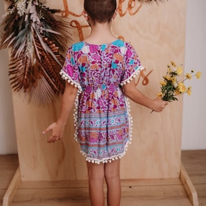 Peut inclure: Robe colorée à motifs floraux roses, violets et bleus. La robe a une taille élastique et des pompons blancs. La personne tient un bouquet de fleurs jaunes. Le fond est un panneau en bois.