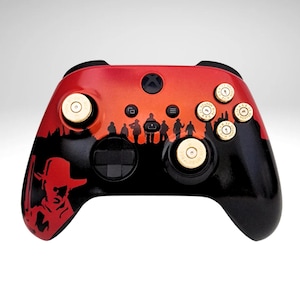 May include: A black and red Xbox controller with custom gold buttons and a silhouette design of a group of people in the centre. The controller has a red and black design with a silhouette of a group of people in the centre. The controller has custom gold buttons.