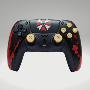 Könnte beinhalten: Schwarzer und roter PlayStation 5-Controller mit einem Spritzer-Design. Der Controller verfügt über ein rot-weißes Regenschirm-Logo und goldfarbene knopfförmige Knöpfe. Der Controller hat ein schwarzes Steuerkreuz und Tasten.