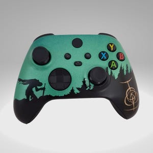 May include: A green and black Xbox controller with a silhouette of a person holding a sword in a forest scene. The controller has a black and gold design on the right side.