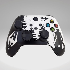 Könnte beinhalten: Ein schwarz-weißer Xbox-Controller mit einem benutzerdefinierten Design, das eine Silhouette eines Waldes, eines Drachen und eines Kriegers zeigt. Der Controller hat ein schwarz-weißes Farbschema mit weißem Hintergrund und schwarzen Akzenten.