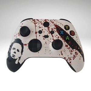 May include: White Xbox controller featuring a black and white graphic of Michael Myers from the Halloween films. The controller has red blood splatter and a black knife graphic.