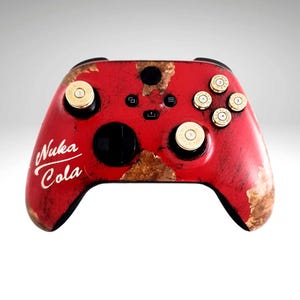 May include: A red Xbox controller with a distressed, weathered look. The controller has gold bullet-shaped buttons and a white sticker that says "Nuka Cola".