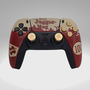 May include: A custom-designed PlayStation 5 controller with a red and beige camouflage pattern. The controller features a custom design with the text "Drink Jugger-Nog" and a 10¢ logo. The controller has gold-colored buttons and triggers.
