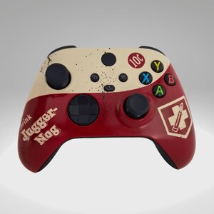 May include: A red and white Xbox controller with a distressed paint design. The controller has a black d-pad, black buttons, and a black faceplate. The controller has the words "Drink Jugger-Nog" and "10¢" printed on it.