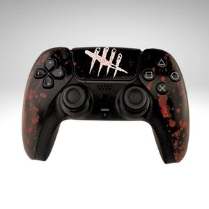 May include: A black PlayStation 5 DualSense controller with a red and black splatter paint design. The controller has a white sticker with the number "4" repeated five times on the top.