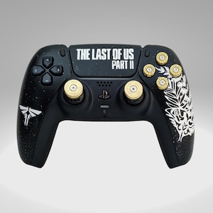 May include: A black PlayStation 5 controller with a white "The Last of Us Part II" logo and a white firefly design. The controller has gold bullet-shaped buttons.