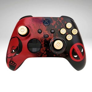 May include: A black and red Xbox controller with a Deadpool design. The controller has gold bullet-shaped buttons and a red and black splatter paint design.