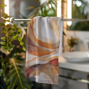 May include: A hanging bath towel with an abstract design. The towel features a flowing pattern of warm colors, including shades of brown, orange, and white. The design resembles watercolor or marble. The towel is hanging on a silver bar in a bathroom setting.